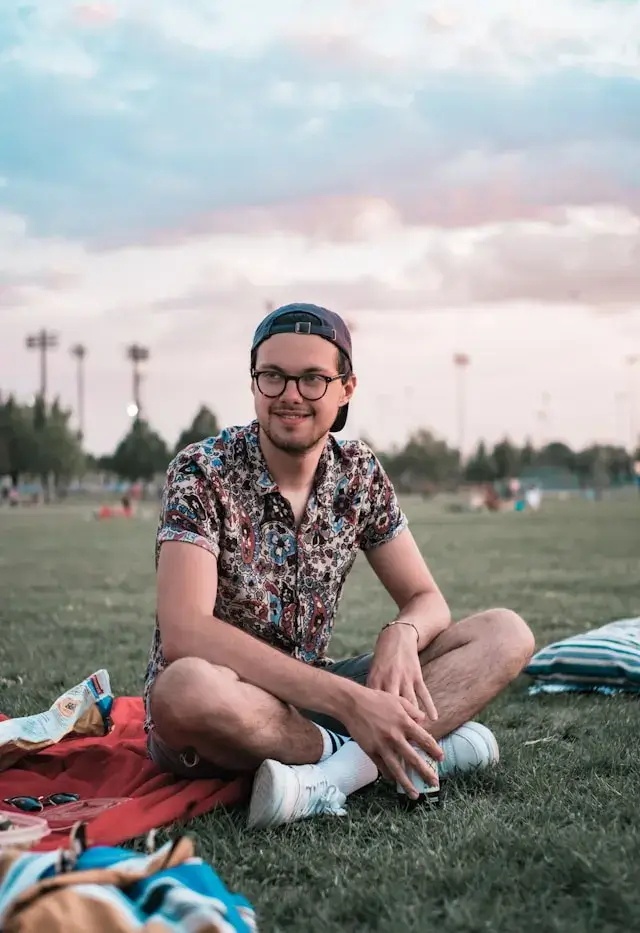 Jeune homme souriant avec lunettes et casquette, portant une chemise à motifs floraux, assis en tailleur sur l'herbe d'un parc avec une couverture de pique-nique rouge, des snacks et une bouteille, sous un ciel nuageux aux tons roses au crépuscule.